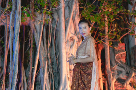 Woman in Thai traditional dress costume, Asian woman wearing Thai dress walking around Ancient City at night Banyan tree background.の写真素材