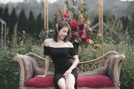 Woman wearing black dress sitting on the couch on flowers garden.