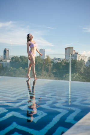 Woman wearing purple bikini standing on the edge of pool on rooftop with city view.の写真素材