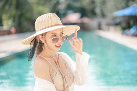 Woman wearing white shirt, long skirt and straw hat posing near swimming pool.