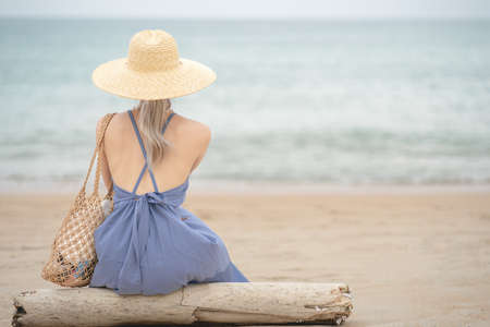 Woman in  blue dress and straw hat, sitting on a timber by the ocean.の写真素材