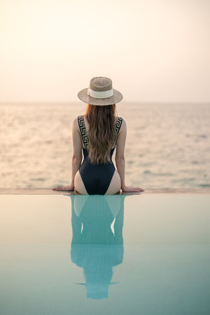 Woman in black swimsuit relaxing by infinity pool with ocean view during sunset.の写真素材