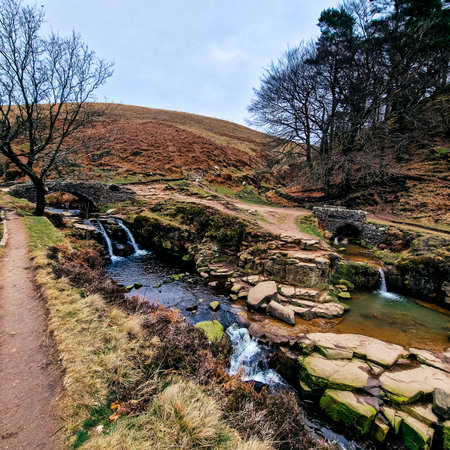 Three Shire Heads, where water cascades over mossy rocks and a packhorse bridge arches over the stream. Lush, rolling hills of the Peak District embrace this enchanting meeting pointの写真素材
