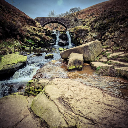 Three Shire Heads, where water cascades over mossy rocks and a packhorse bridge arches over the stream. Lush, rolling hills of the Peak District embrace this enchanting meeting pointの写真素材