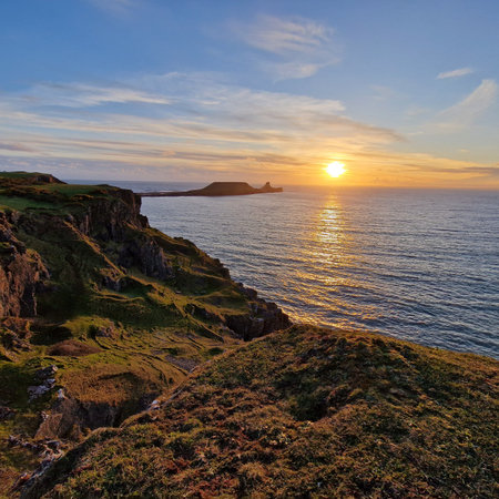 In this breathtaking photograph, the sun sets behind the iconic Worm's Head, casting a golden glow...
