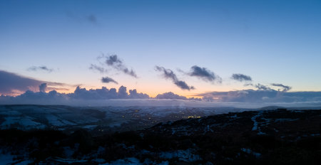 The photograph captures view from Bamford Edge during winter after the sunset. The sky is adorned with hues of blue and purple, creating a serene and wintery atmosphere. The snow-covered landscape glistens under the fading light, painting a peaceful and tranquil scene. The silhouettes of trees and rocky formations stand tall against the wintry backdrop, adding a touch of mystery and beauty. It's a moment where nature embraces the stillness and quietude of the season, creating a captivating and enchanting winter landscape at Bamford Edge.の写真素材