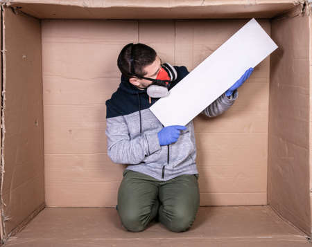 man in a protective mask is sitting in a cardboard box, holding an empty board with a place for an advertisement,の写真素材