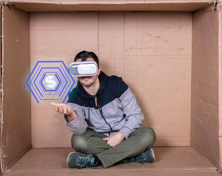 man with glasses for virtual reality sits in a cardboard room and shows a dollar symbolの写真素材