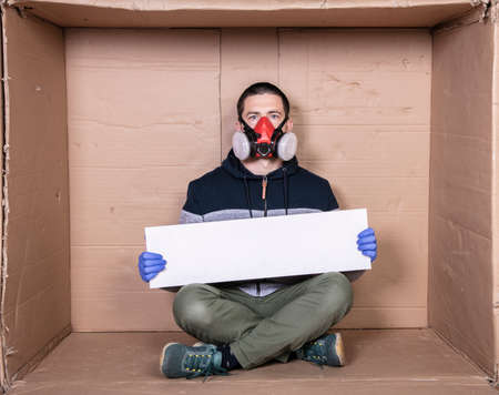 man in a protective mask sits at a cardboard box, holds an empty sign with a place for an...
