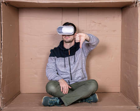 man with glasses for virtual reality, sitting in a cardboard room and showing thumbs downの写真素材