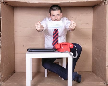 businessman showing a protective mask, sitting in his little cardboard officeの写真素材