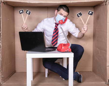 businessman works in an uncomfortable cardboard office, quietly calls for help, in the meantime talking on the phoneの写真素材