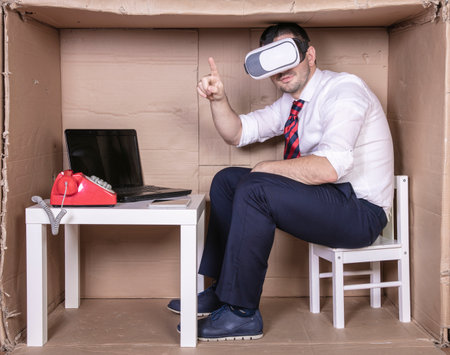 businessman sitting in a cramped cardboard office, virtual glasses on his head, finger in the air, copy spaceの写真素材