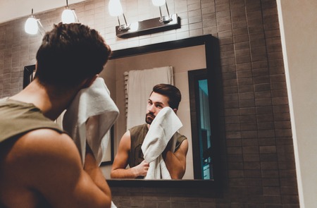 Reflection of young man in bathroom mirror looking on his faceの写真素材
