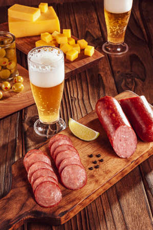 Sliced smoked salami on cutting board with two glasses of beer, cubs of cheese and olivesの写真素材