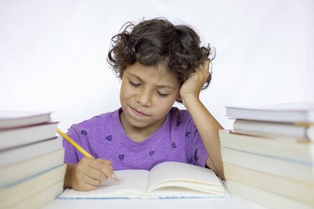 Happy boy between 8 and 10 years old with expression of doubt sitting at a desk doing his homework,...