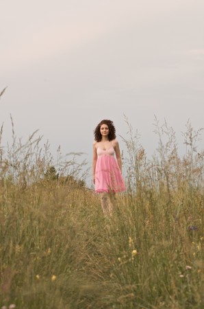 A young woman wearing a pink dress is smiling and standing in a field. Vertical shot.の写真素材