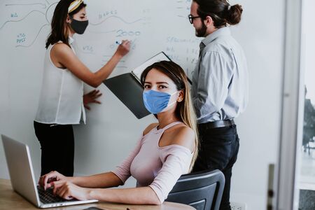 Latin woman working in business office or coworking while wearing medical face mask for social distancing in new normal situation protecting and preventing the infection of covid-19, mexican coworkers in Mexico cityの写真素材