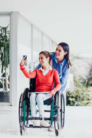 Young latin woman in wheelchair taking a selfie photo at workplace with another woman in Mexicoの写真素材