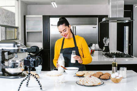 Latin Woman baking Conchas traditional Mexican bread while recording a video class with camera in a kitchen in Mexico cityの写真素材