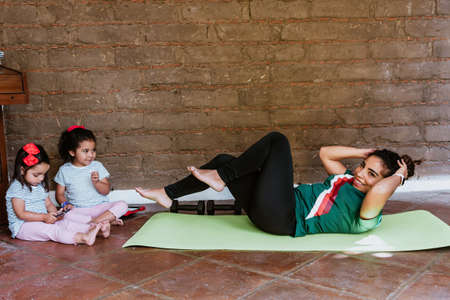 Sporty latin woman mother in sportswear is exercise on the floor with daughter at home in Mexico...