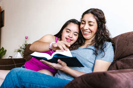 Smiling latin teen girl with down syndrome and her mom reading a book at home, in disability concept in Latin Americaの写真素材