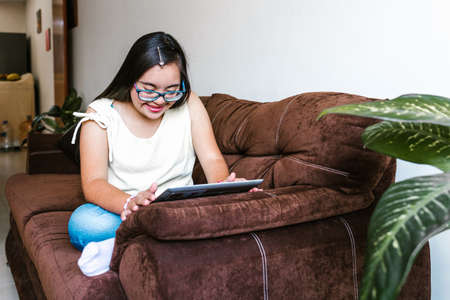 Hispanic teen girl with down syndrome using a tablet while sitting on the sofa at home, in disability concept in Latin Americaの写真素材