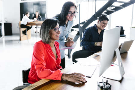 Mexican transgender woman sitting in wheelchair and office colleagues working on project using computer in office in Latin Americaの写真素材