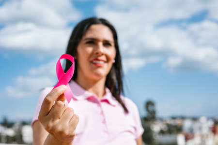 portrait of latin woman hand holding pink breast cancer awareness ribbons with blue sky as...