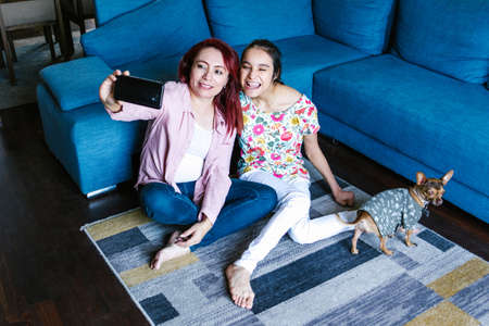 Latin teen girl with cerebral palsy and her mother taking selfie sitting in the living room in...