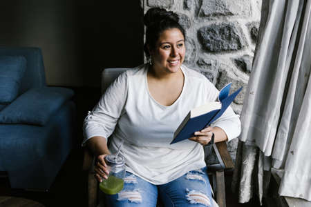Portrait of curvy latin girl holding a jar of green juice while reading a book at homeの写真素材