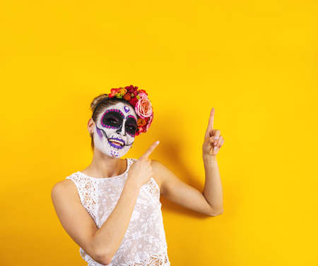 Mexican Catrina, portrait of young latin woman pointing aside on yellow copy space, Halloween party...