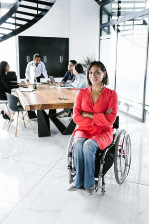 Portrait of latin transgender woman in wheelchair and people at meeting in the background in the...