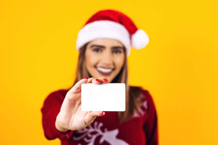 Portrait of Latin woman holding an empty blank card for shopping at Christmas wearing a santa hat on yellow background in Mexico Latin Americaの写真素材