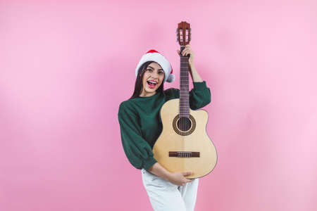 Portrait of young latin woman holding guitar music with copy space in a christmas concept on pink backgroundの写真素材