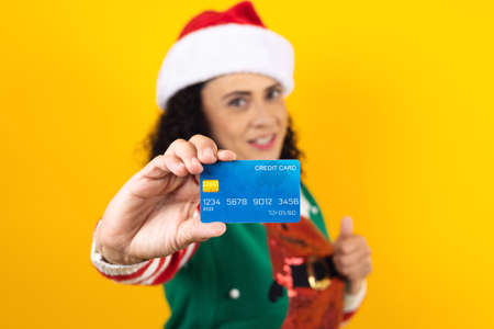 Portrait of adult woman holding a credit card for shopping at Christmas wearing a santa hat on yellow background in Mexico Latin Americaの写真素材