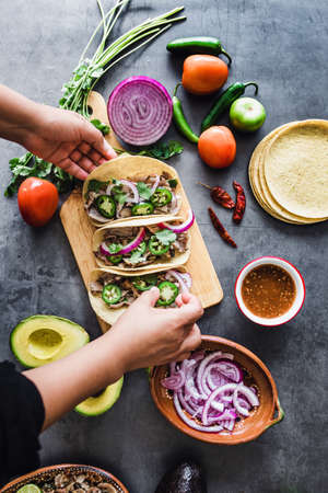 latin woman hands preparing mexican tacos with pork carnitas, avocado, onion, cilantro, and red sauce in Mexicoの写真素材