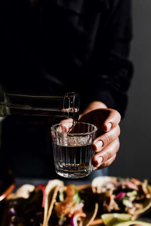 Barman hands serving mexican mezcal shot in a traditional glass with tacos and food at background in Mexicoの写真素材