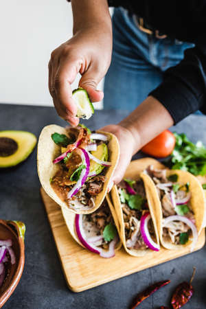 latin woman hands preparing mexican tacos with pork carnitas, avocado, onion, cilantro, and red sauce in Mexicoの写真素材