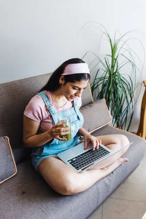 young latin Woman student using laptop in a video call or online class and drinking green juice while sitting on sofa at home in Mexicoの写真素材