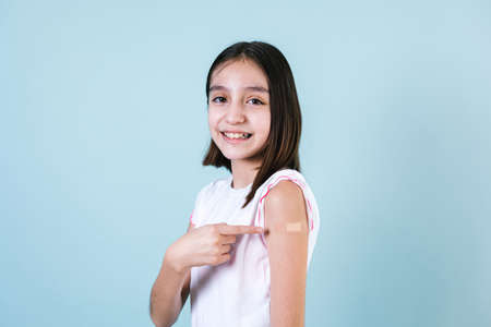 Hispanic girl child portrait after getting a vaccine protection and showing her arm with bandage receiving vaccination on blue background in Mexico Latin Americaの写真素材