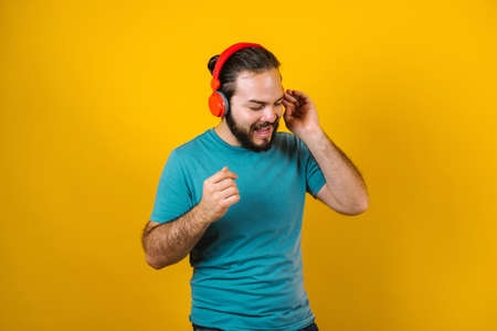portrait of hispanic young man with headphones, dancing and listening music on yellow background in...