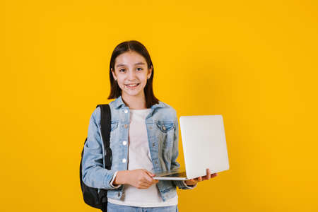 portrait of young hispanic child teen girl student with computer lap top on yellow background in...