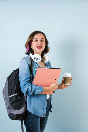 Young Hispanic student woman wearing backpack and holding books and coffee cup over isolated blue...