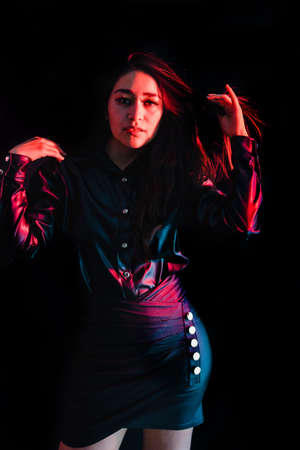 young latin woman dressed in black clothes with colorful pink-blue lights on black background in...