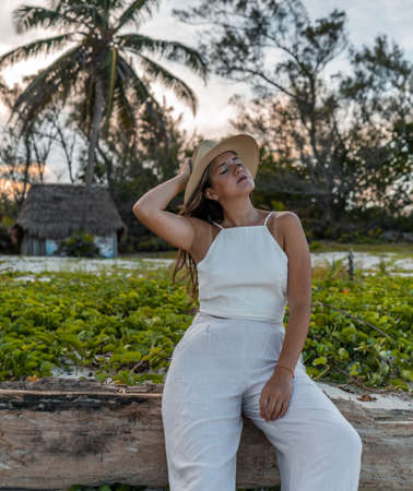 Young model with hat on the beach sitting on a wooden log with her eyes closed - enjoying fresh airの写真素材