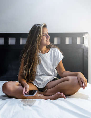 young hispanic woman sitting on the bed smiling with cell phone and headphones looking to her left listening to her favorite musicの写真素材