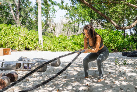 young hispanic woman does arm exercises with ropes in a beach eco gymの写真素材