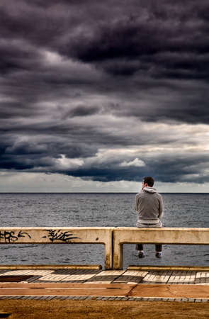 Man sitting on a pier and looking at a stormy sky.の写真素材