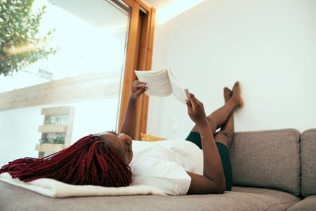 Black woman laying on the sofa reading a book with his feets on the wallの写真素材